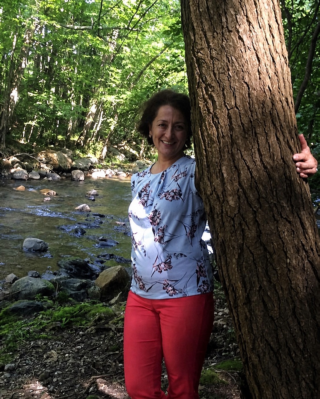 Gulnara Karimova standing beside a large tree and a woodland creek.